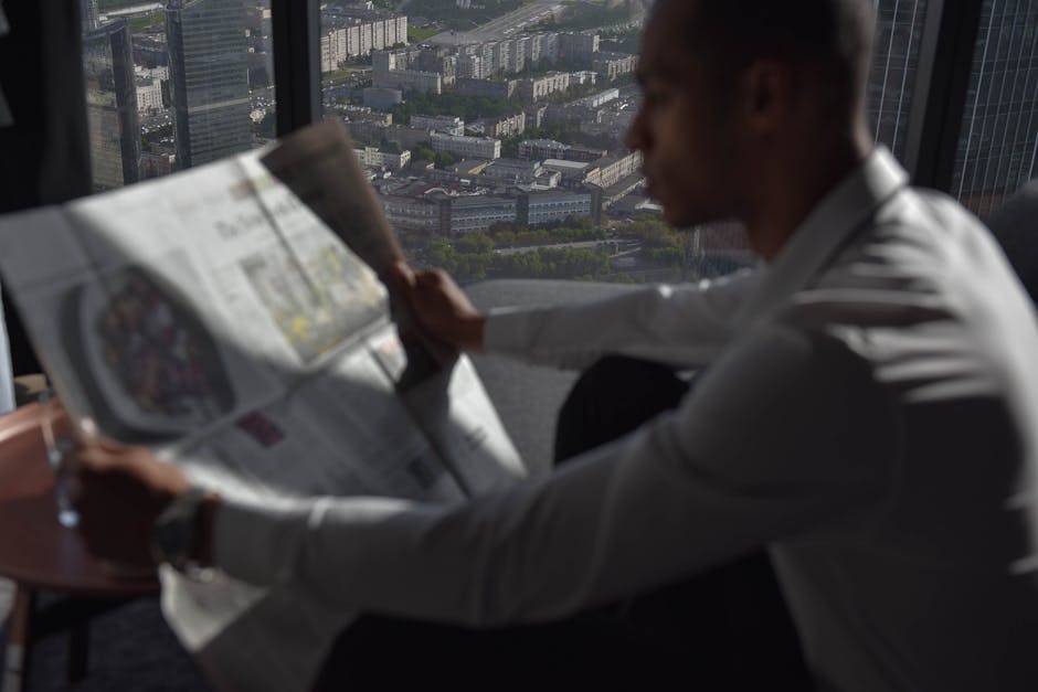A man in a white shirt reads a newspaper with a high-rise city view.