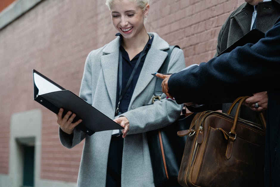Low angle of crop happy young female employee in formal clothes smiling while reading documents standing near brick building with faceless colleagues